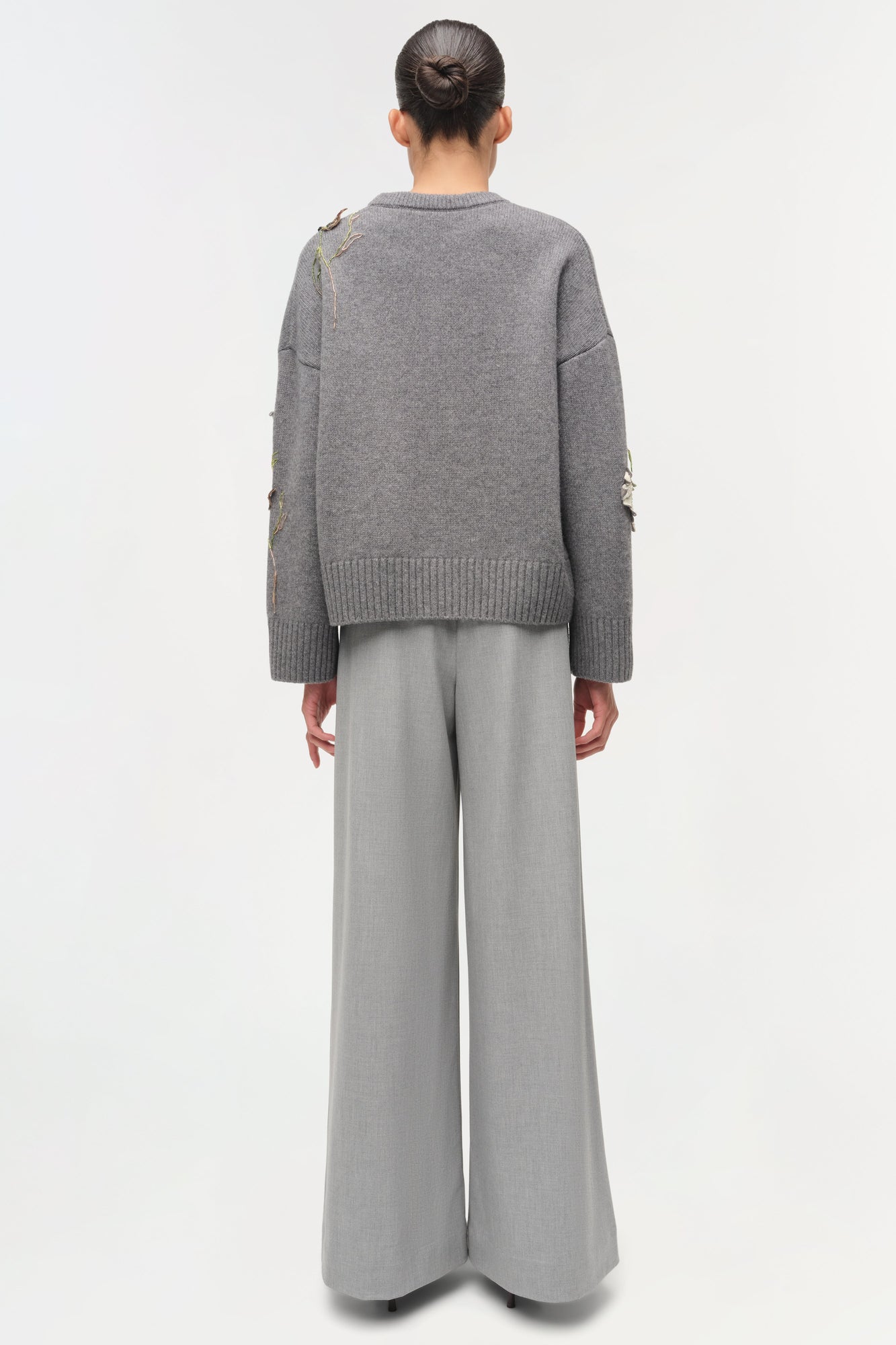 Gray knit sweater with floral embellishments on sleeves, worn with light gray wide-leg pants, back view.