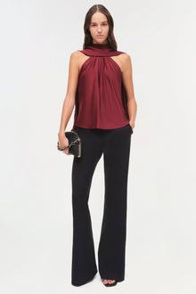 Woman wearing burgundy sleeveless halter top with draped cape detail and black flared pants.
