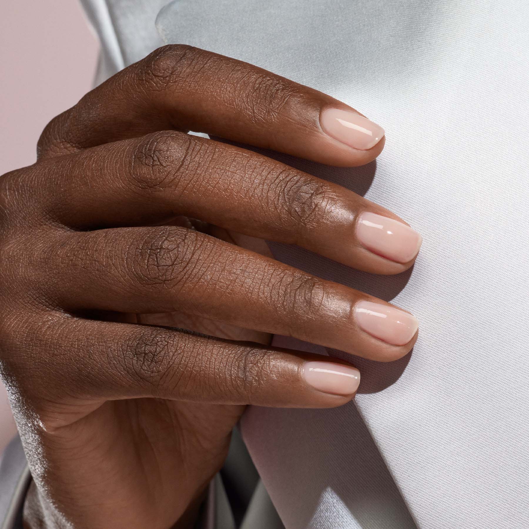 A hand with short, neatly manicured nails painted in Manucurist’s Pale Rose, a sheer pastel pink polish, rests gently on white fabric.