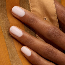 A close-up of a hand with medium brown skin shows neatly manicured nails painted in Manucurist's Nebula, a shimmery milky white polish, resting on gold and golden brown striped fabric.