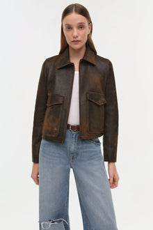 Brown leather jacket with spread collar, front zip closure, and two flap chest pockets worn over white tee and light blue jea