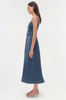 Woman wearing a blue denim sleeveless dress with a belted waist, shown from the side profile.