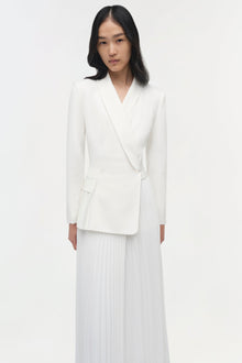 Woman wearing a cream blazer dress with pleated skirt detail and long sleeves.