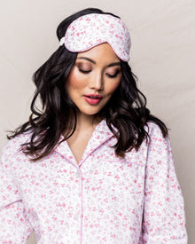 A woman with long, dark hair wears Petite Plumes Adults Sleep Mask in Dorset Floral, matching her pink floral pajama set. She gazes downward, standing against a light textured background—a perfect gift for those who appreciate comfort and style.