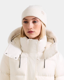 A woman wears the Rudsak ANDREY - U425507 Vanilla rib-knit hat and thick white puffer jacket with high collar and hood, looking slightly to the side against a plain light background.