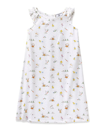 A young girl with short brown hair smiles in the Girl's Twill Amelie Nightgown in Easter Frolic, a white sleeveless dress featuring playful animal and nature prints, standing against a plain light background.
