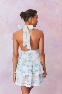 White halter dress with a blue floral pattern, halter ties in bow at neck and below, is a wide smocked waistband and the skirt has two ruffled tiers.