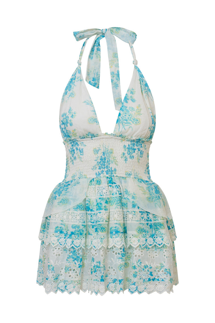 White halter dress with a blue floral pattern, halter ties in bow at neck and below, is a wide smocked waistband and the skirt has two ruffled tiers.