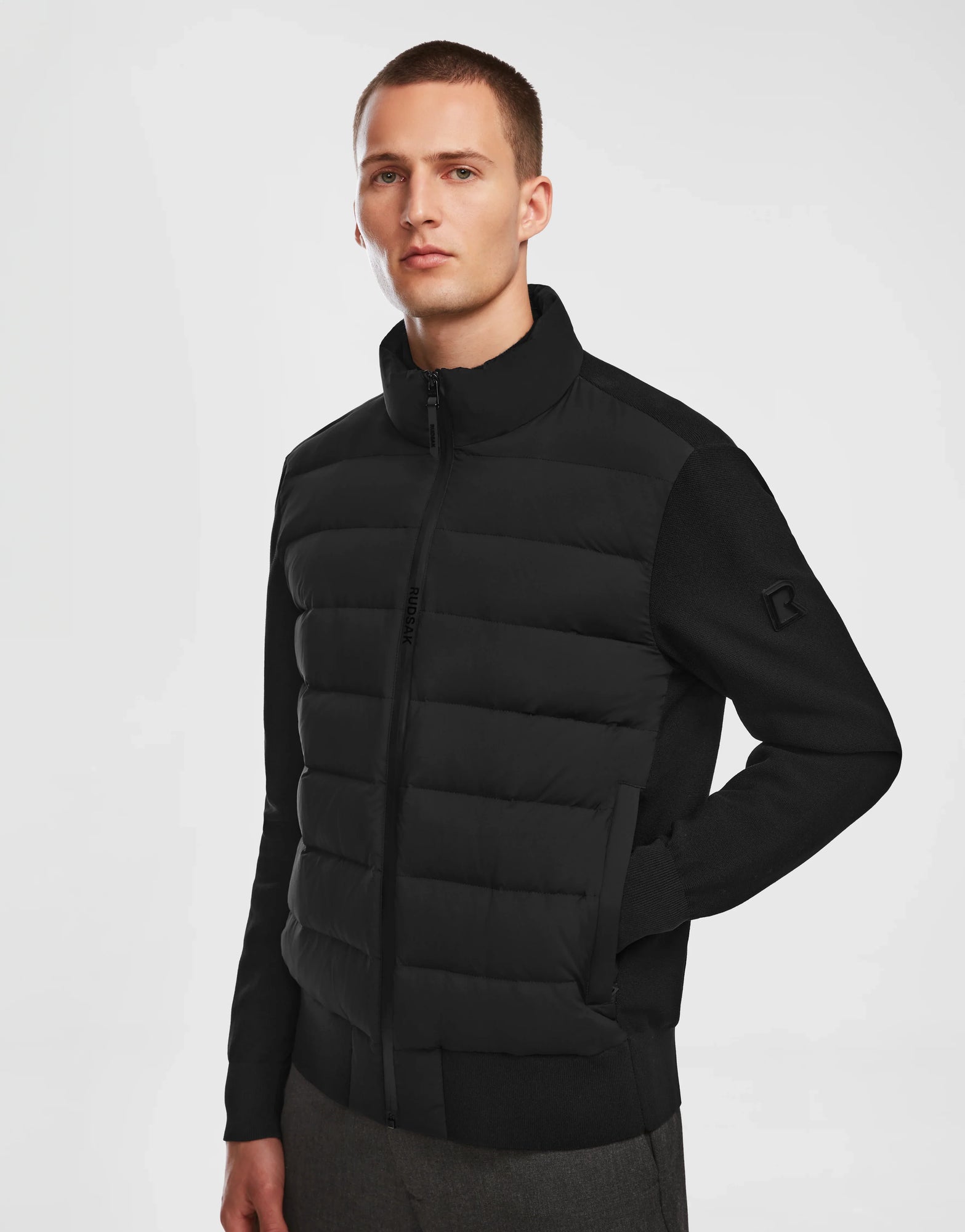 A young man with short hair wears the Rudsak EUGENE - 6124016 Black mens hybrid jacket, hands in pockets, standing against a plain white background.
