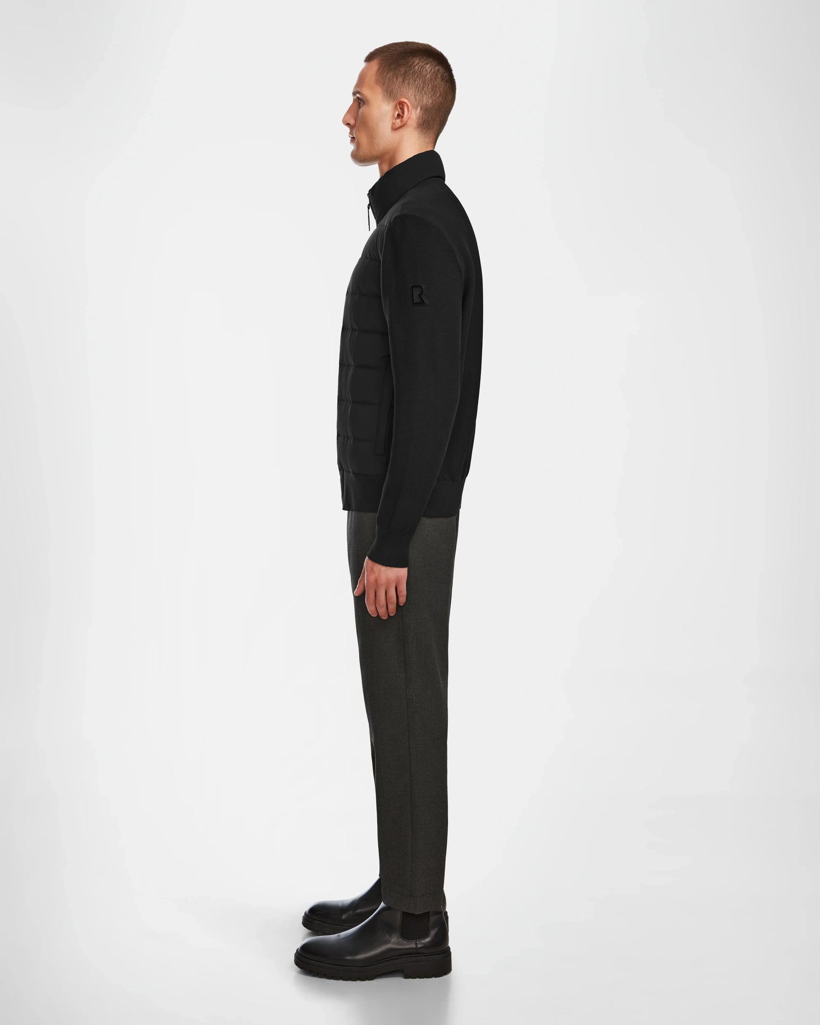 A man stands in profile against a plain white background, wearing the Rudsak EUGENE - 6124016 Black mens hybrid jacket, paired with dark trousers and black boots.