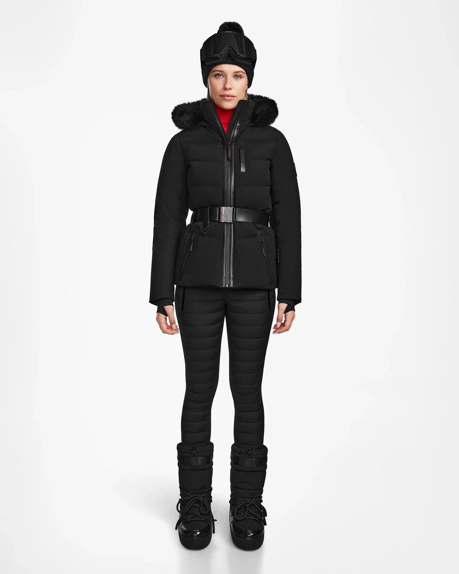 A person stands facing forward, wearing the Rudsak ALAIA - 8124816 Black women’s quilted ski jacket with a fur-trimmed hood, matching pants, winter boots, wide belt, black hat, and RECCO rescue technology for cold weather safety.