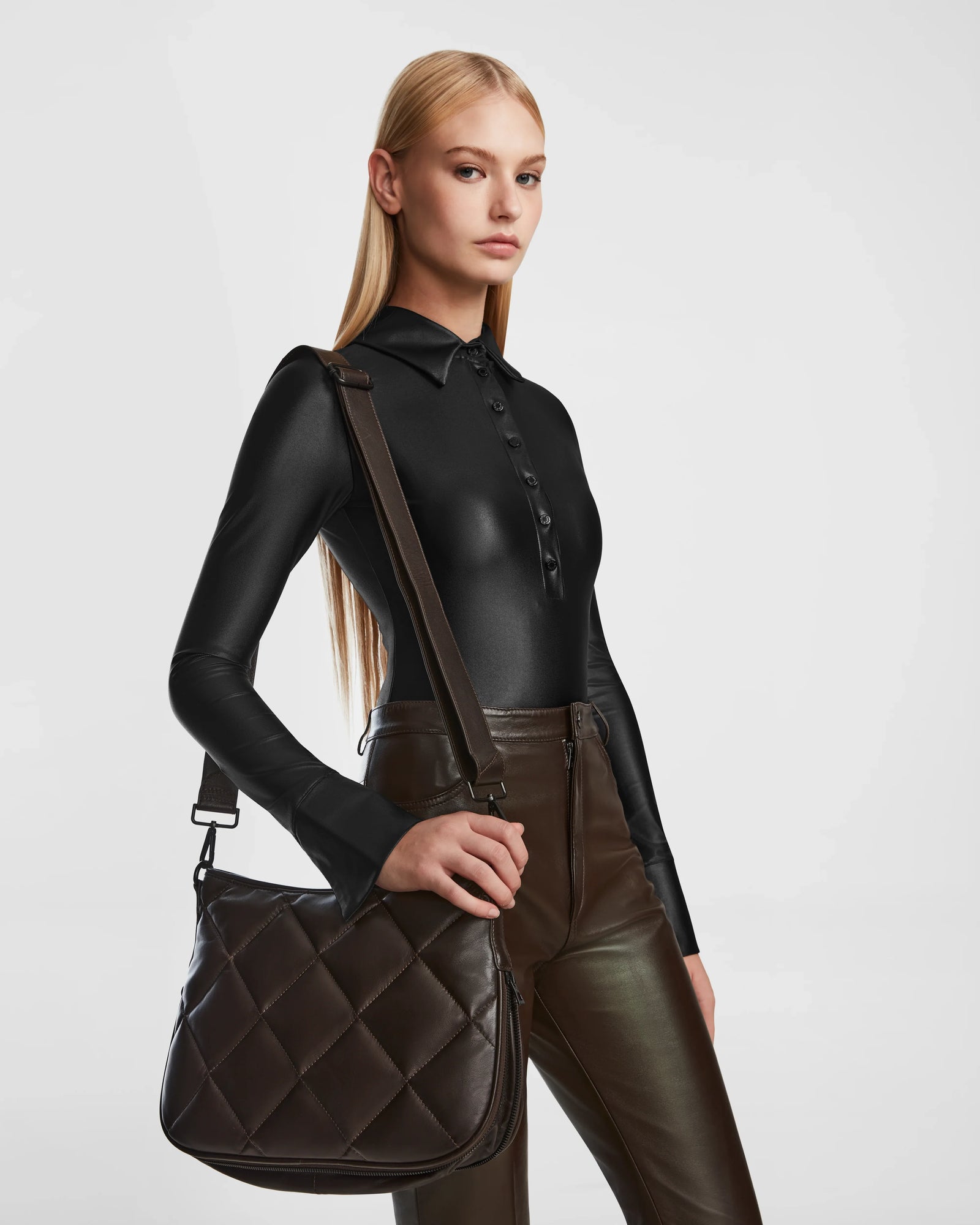 A woman with straight blonde hair wears a fitted black long-sleeve top and brown leather pants, holding the Rudsak ALICIA - 8323507 Espresso luxe quilted leather purse with a long strap against a plain light background.