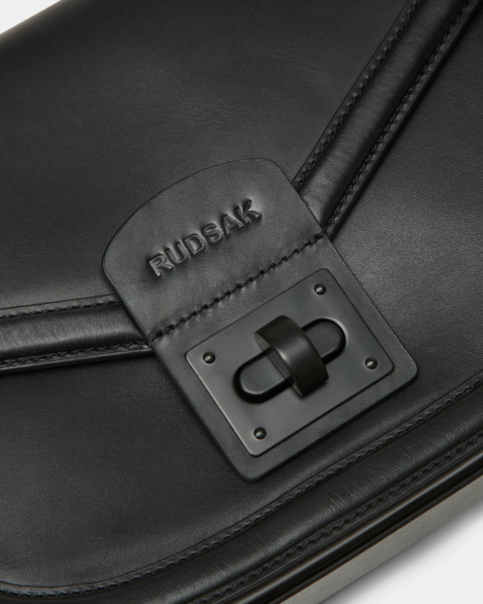 Close-up of Rudsaks ALORA - 8324506 Black womens leather crossbody bag, showcasing a logo-embossed flap above a rectangular black clasp for a sleek, premium look.