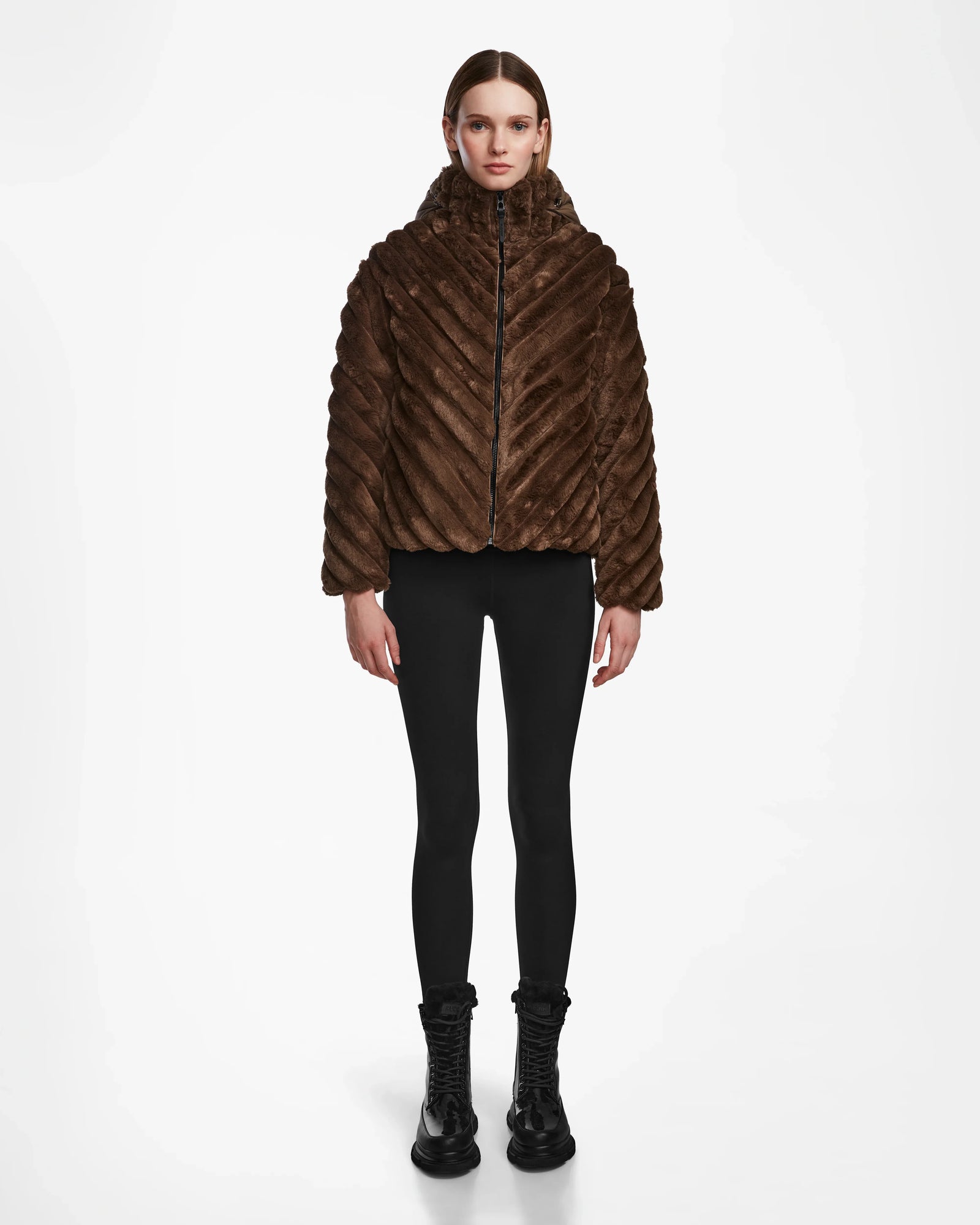 A person faces forward, wearing the Rudsak ANASTASIA - 8124804 Espresso dark brown winter jacket, paired with black leggings and black lace-up boots, set against a plain white background.