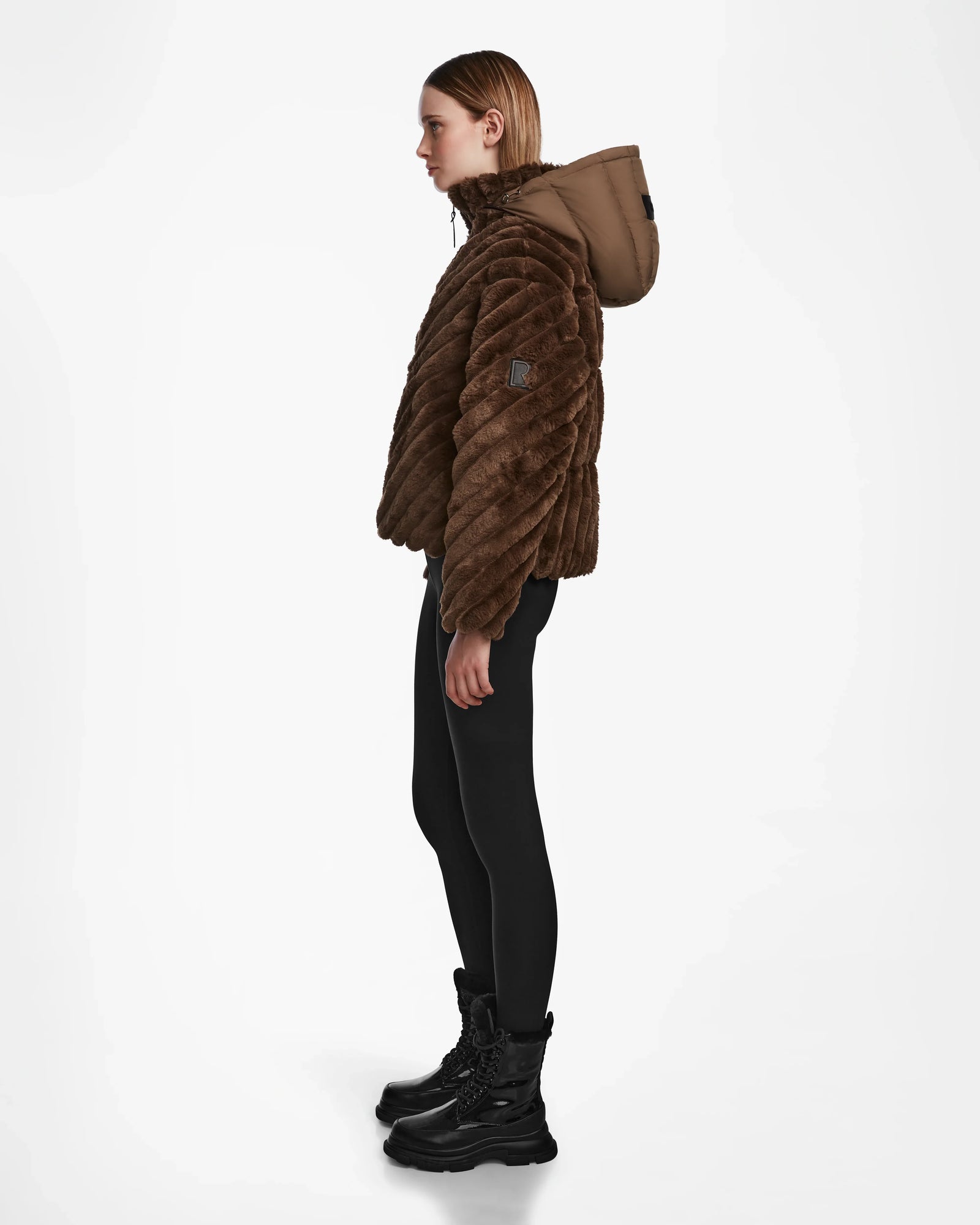 A person stands in profile against a plain white background, wearing the Rudsak ANASTASIA - 8124804 Espresso brown velvet puffer hooded jacket, black leggings, and black lace-up boots.