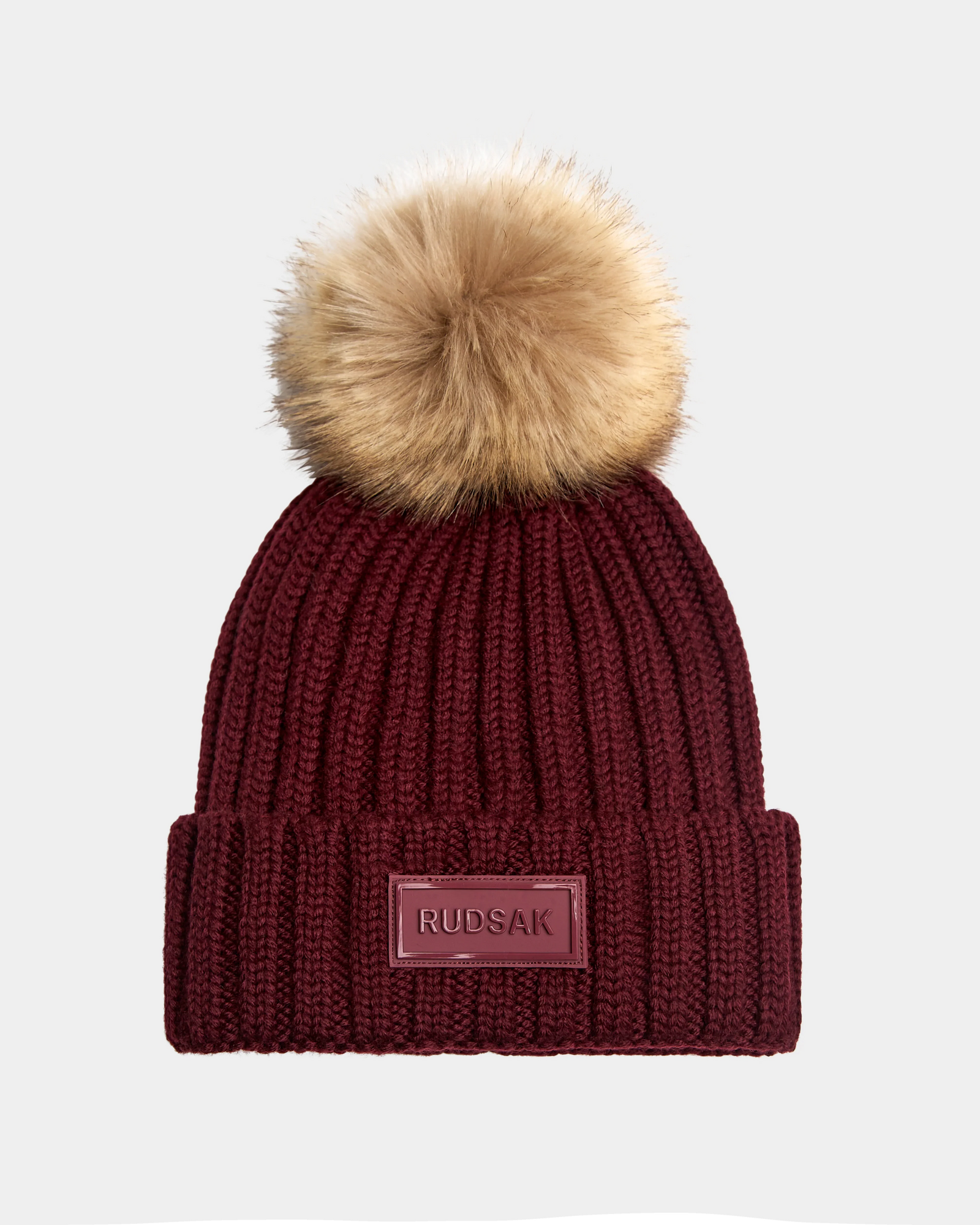 The ANISA - L423500 Wine by Rudsak is a maroon ribbed knit women’s beanie with a wide folded brim, brown faux fur pom pom, and a front rectangular Rudsak patch—perfect for adding style to your winter look.