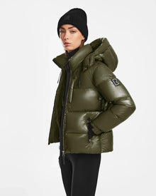 A person wearing a Rudsak ASHLYN S - 8124565 Loden Green womens down puffer jacket with hood and B patch on the sleeve, plus a black beanie, stands against a plain white background. The jacket is RDS-certified.