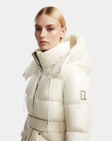 A blonde woman in a sleek low part wears the off-white Rudsak AUDREY L - 8124585 Iceland knee-length down puffer with high collar, hood, RDS certified down, and B logo patch on the sleeve, posing against a plain background.