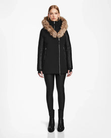 A person faces forward against a white background, wearing the Rudsak AURORE R - L124535 Black women’s down parka with vegan leather sleeves, black leggings, and black boots. The coat features a tan faux fur hood for warmth.