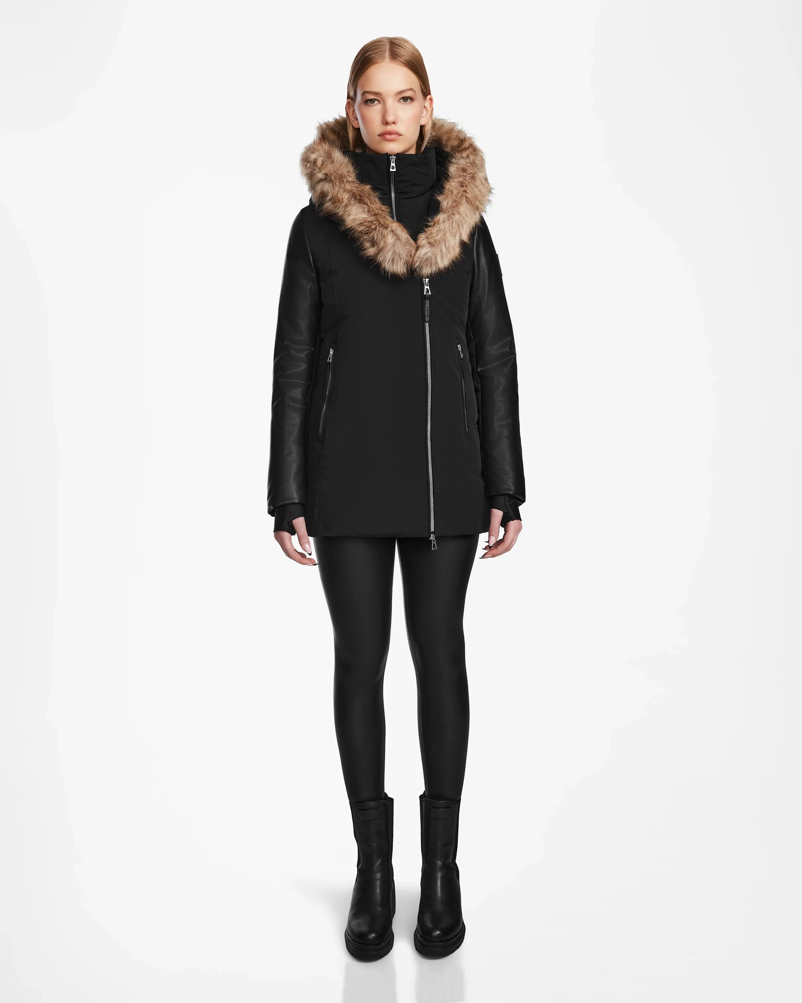 A person faces forward against a white background, wearing the Rudsak AURORE R - L124535 Black women’s down parka with vegan leather sleeves, black leggings, and black boots. The coat features a tan faux fur hood for warmth.
