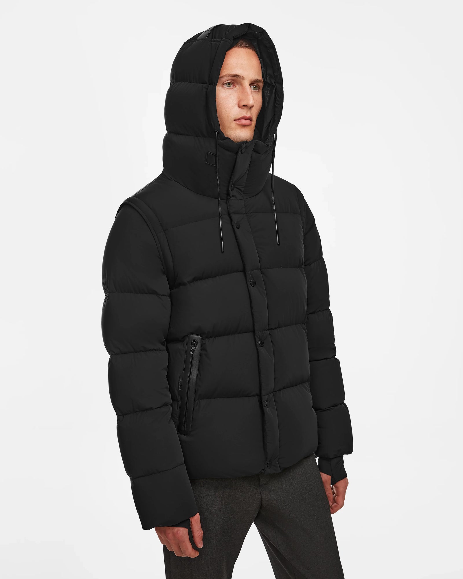 A person stands against a white background wearing the Rudsak BOBBY - 6124506 Black mens down puffer, which is water-resistant, buttoned and zipped with a padded hood, high collar, side pockets, and paired with dark pants.