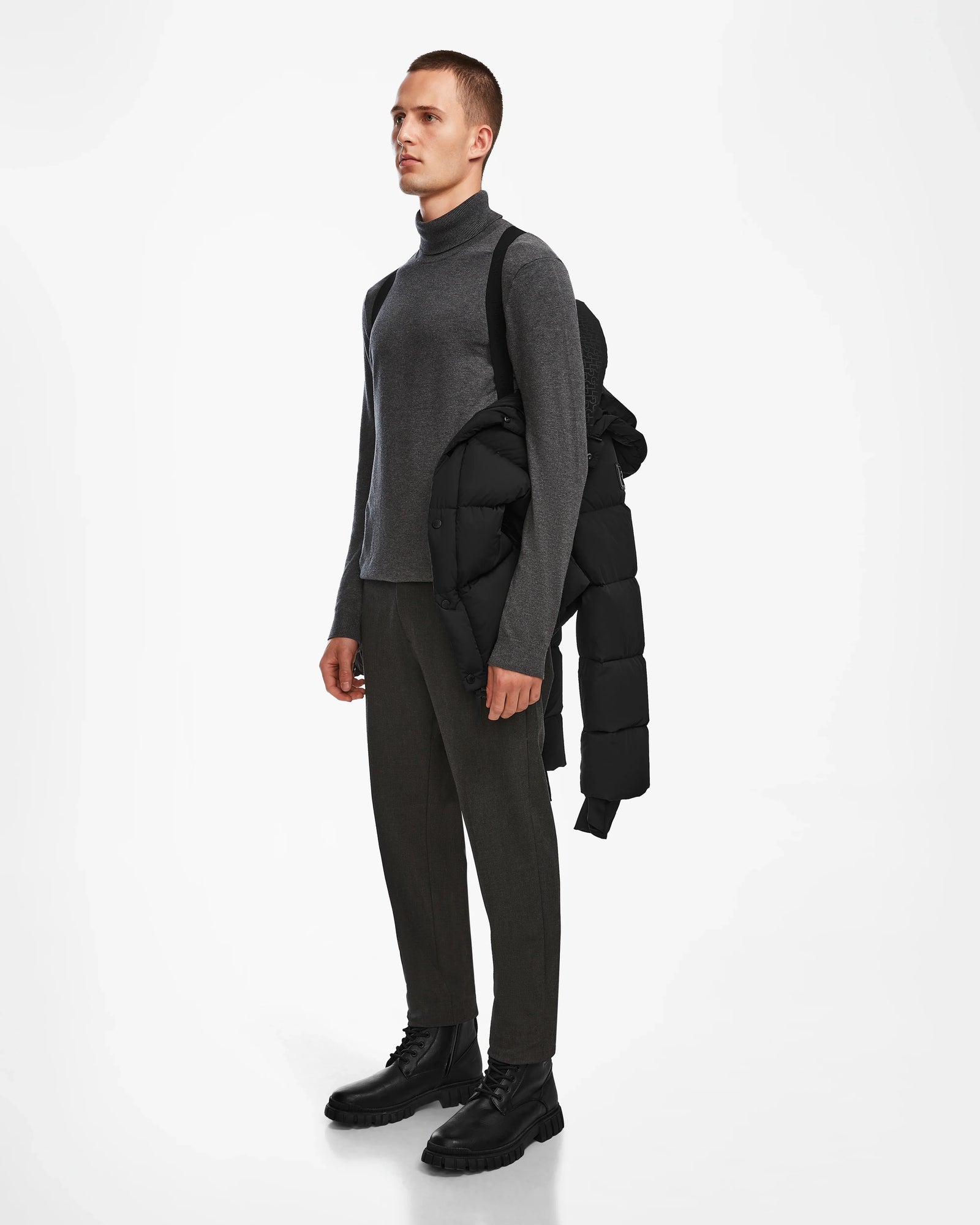 A man stands against a white background wearing a gray turtleneck, dark pants, and black boots, with the Rudsak BOBBY - 6124506 Black men’s down puffer draped over his shoulder.