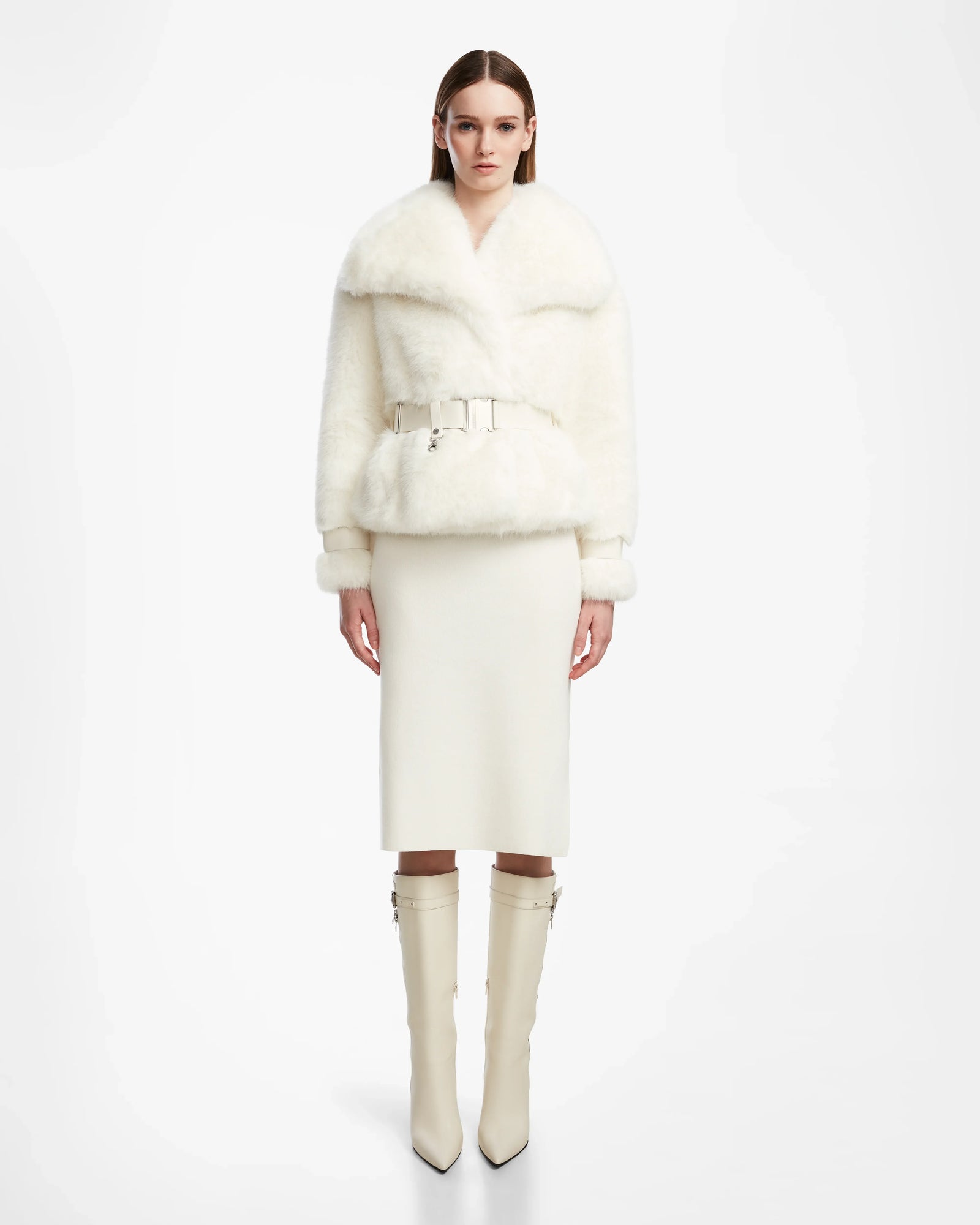 A woman faces forward in the Rudsak CHLOE - L124564 Iceland womens faux fur jacket with a wide collar and belt, styled with a white knee-length skirt and pointed-toe knee-high boots, set against a plain white background.