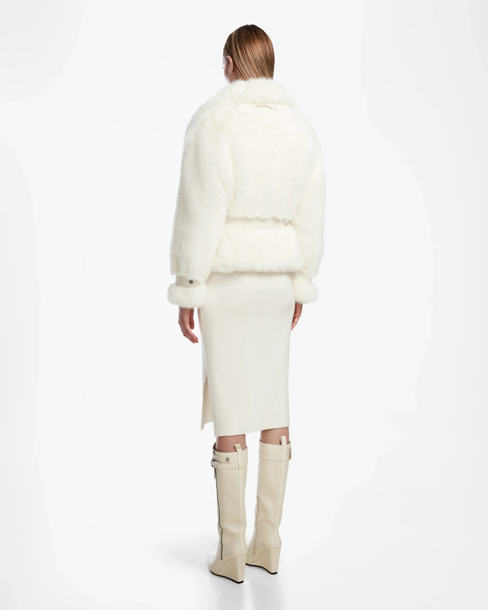 Wearing the Rudsak CHLOE - L124564 Iceland faux fur jacket, a woman with straight, light brown hair pairs it with a white below-the-knee skirt (side slit) and tall cream boots, standing away from the camera—a chic winter wardrobe essential.