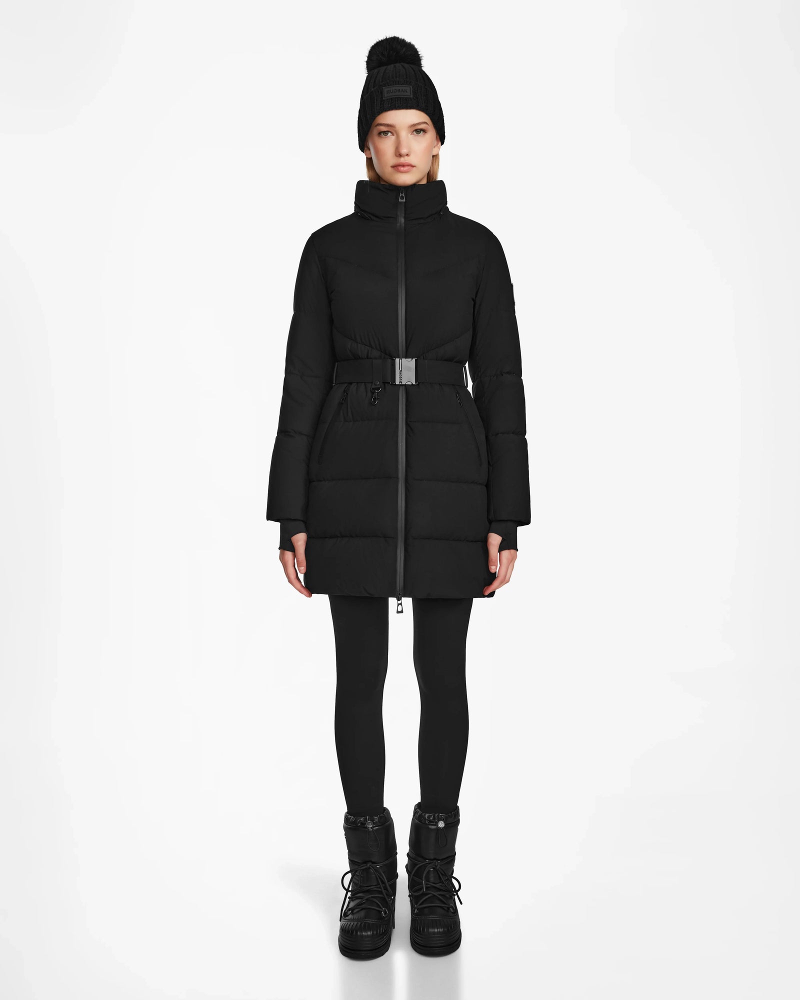 A person wearing the Rudsak CHRISTEL M-M - L124505 Black down puffer with a belt, paired with black leggings, winter boots, and a black pom-pom knit hat, stands against a plain white background.