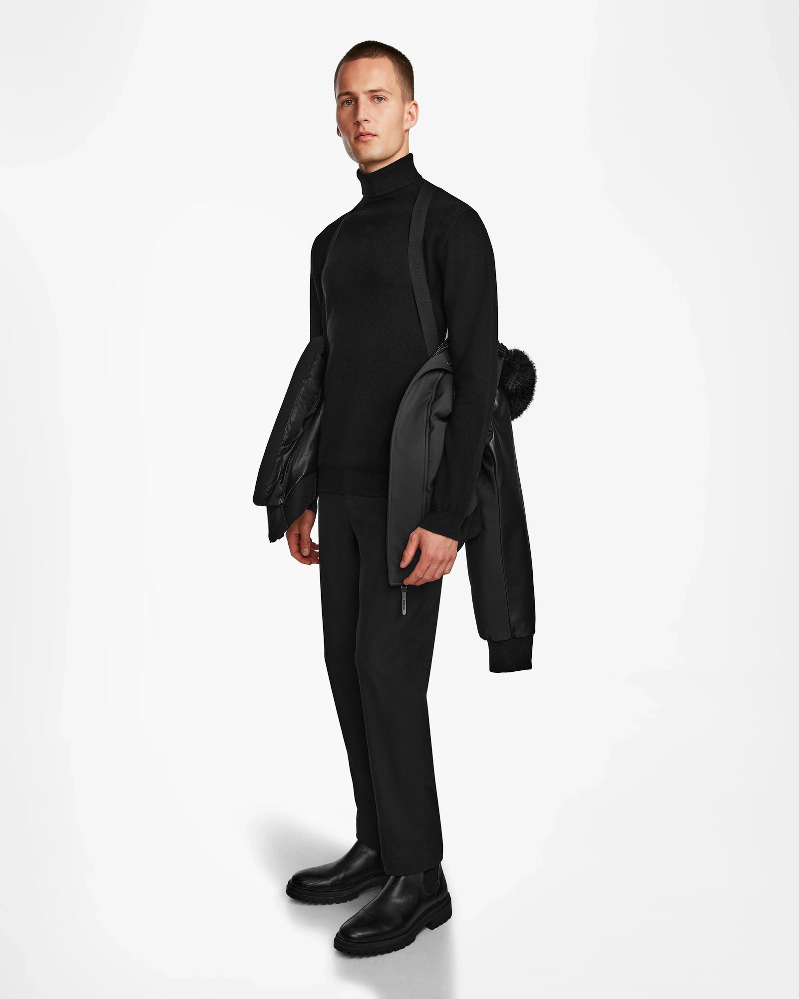 A man stands against a white background wearing black clothes and holds the Rudsak DALLAS R - M123501 BLACK men's quilted down bomber with recycled leather sleeves over his shoulder, looking straight ahead with a neutral expression.