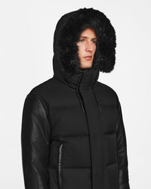 A person wearing the Rudsak DALLAS R - M123501 BLACK: a men's quilted down bomber with recycled leather sleeves and a black fur-lined hood. Its high collar and weather-resistant design stand out against a plain white background.