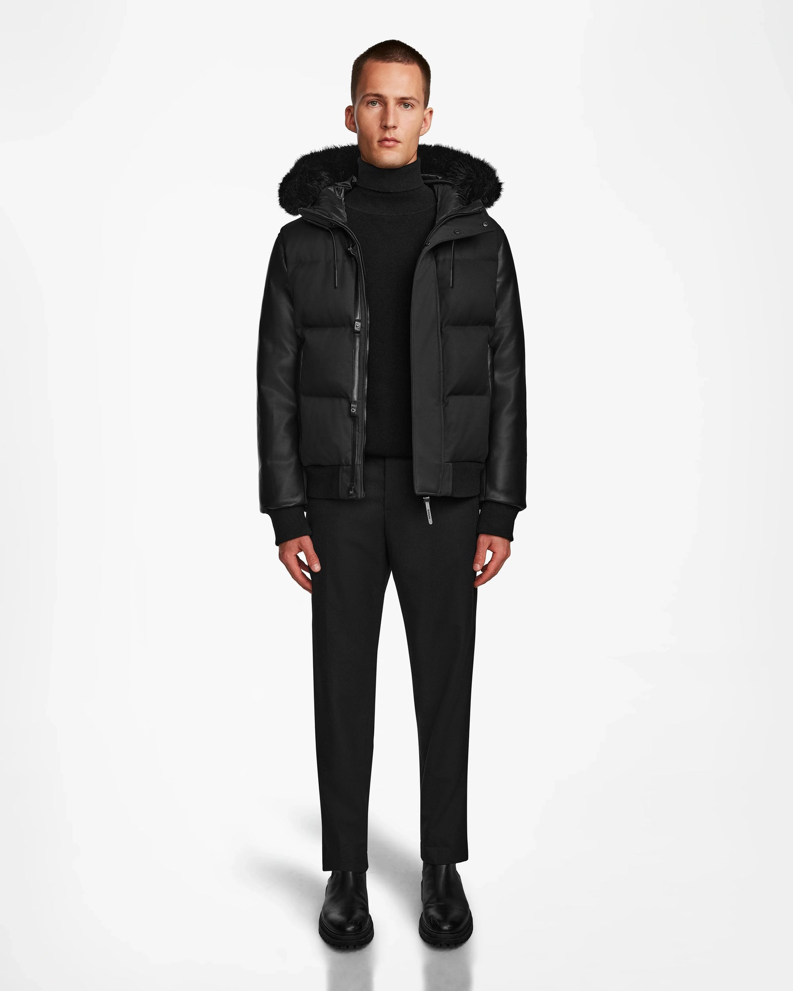 A person faces forward in a Rudsak DALLAS R - M123501 BLACK men's quilted down bomber with recycled leather sleeves, styled with a black turtleneck, pants, and shoes against a plain white background.