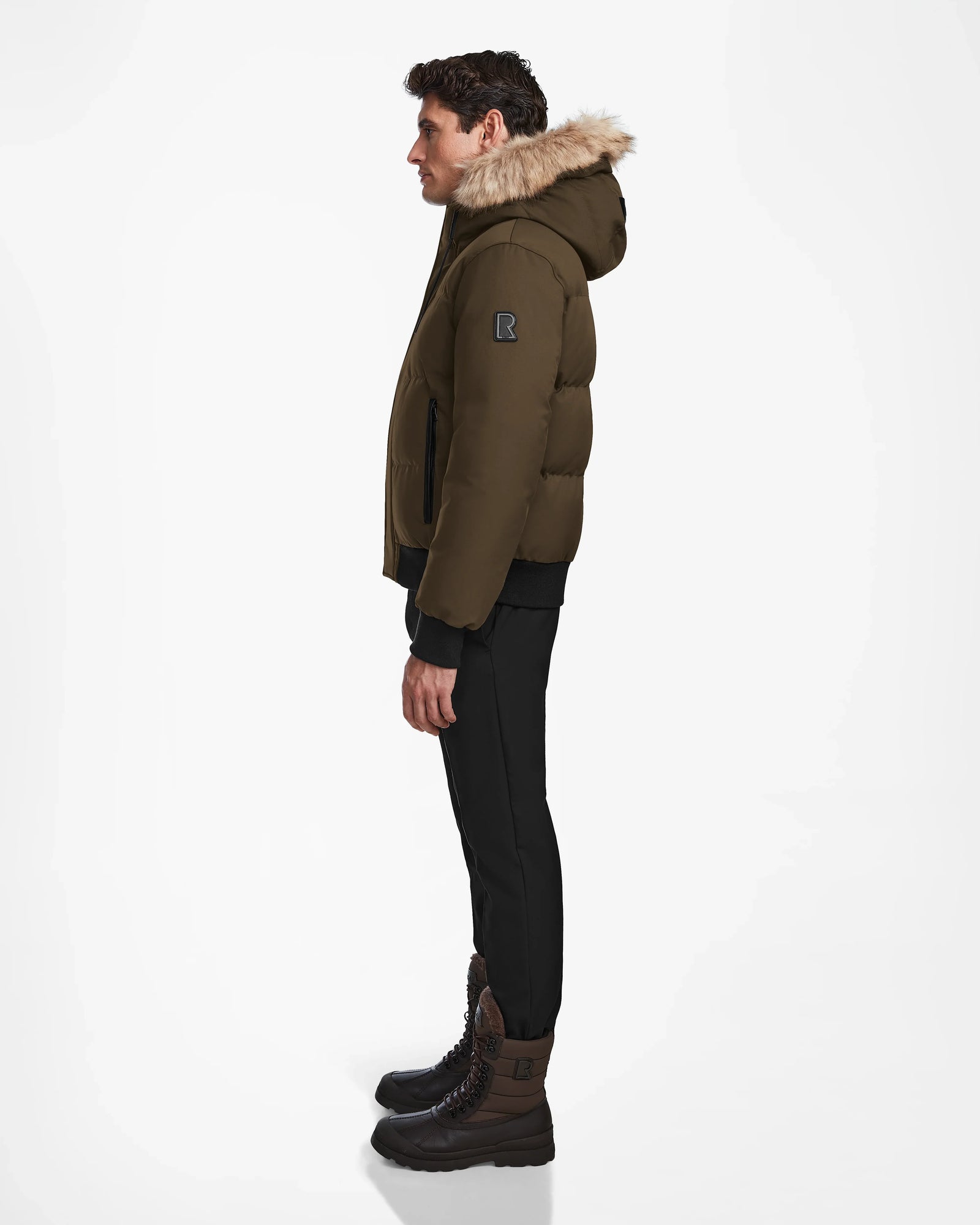 A man stands in profile against a white background, wearing the Rudsak DALLAS - M122501 Army weather-resistant puffer jacket with fur trim, black pants, and brown lace-up boots.