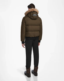 A person faces away, wearing the Rudsak DALLAS - M122501 Army weather-resistant puffer with a fur-lined hood, black pants, and brown boots against a plain white background.