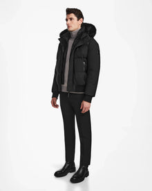 A man stands against a plain white background wearing the Rudsak DALLAS - M122501 Black weather-resistant jacket with fur trim, a light gray turtleneck sweater, black pants, and black leather boots.