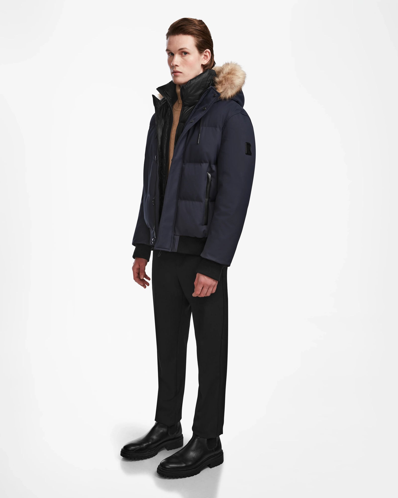 A young man models the Rudsak DALLAS - M122501 Navy men’s quilted down bomber with a fur-trimmed hood, styled with black pants and boots. He stands slightly turned against a white background, highlighting the weather-resistant jacket.