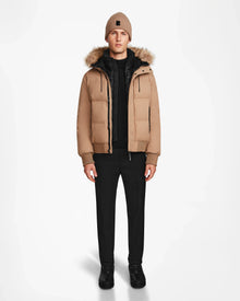 A person faces forward against a white background wearing the Rudsak DALLAS - M122501 Sand weather-resistant bomber jacket with a fur-lined hood, paired with a tan beanie, black pants, and black shoes.