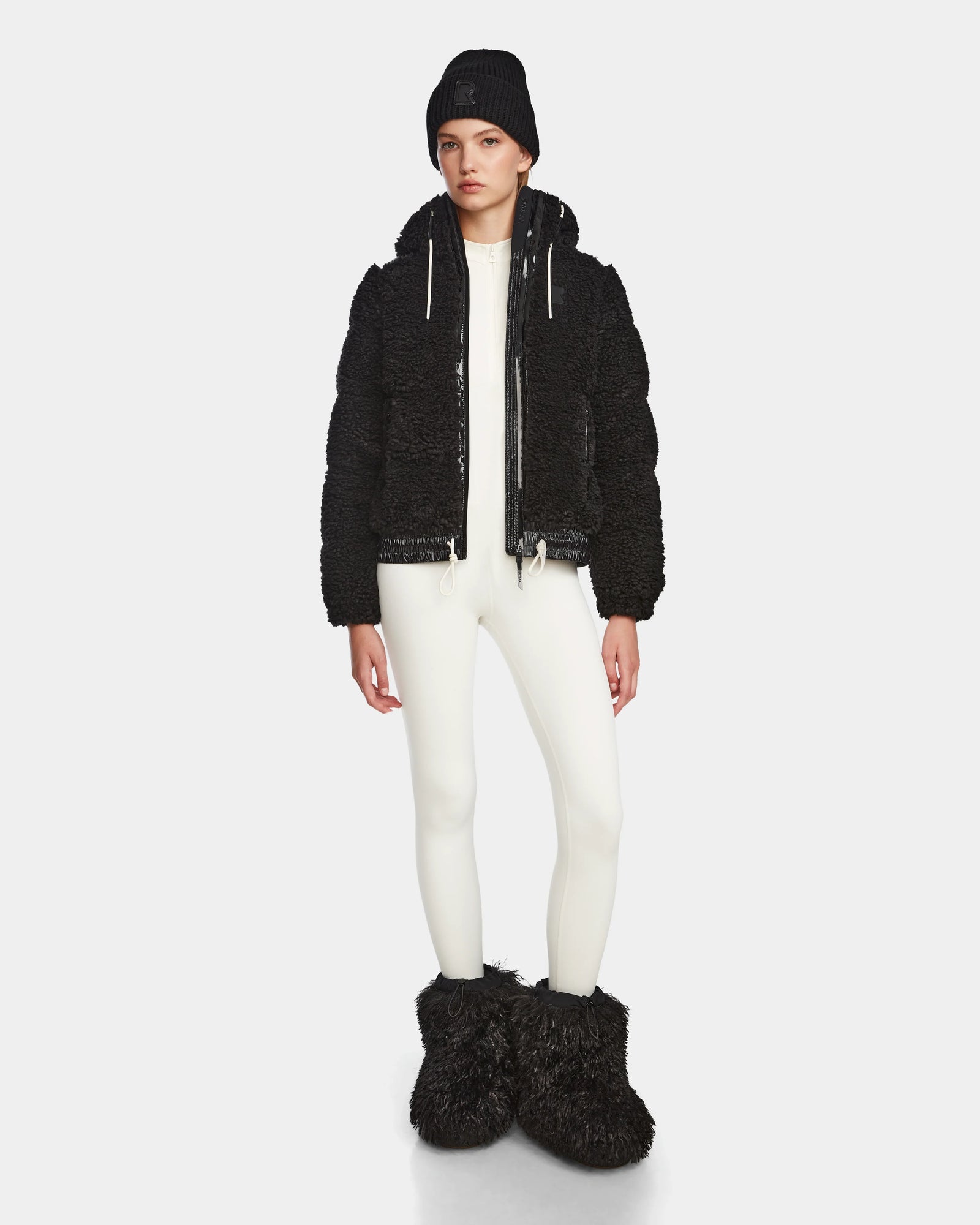 A person stands facing forward in the Rudsak DANI S - 8124537 Black bomber-style ski jacket, paired with a black beanie, white fitted outfit, and large black fuzzy boots against a plain light background.