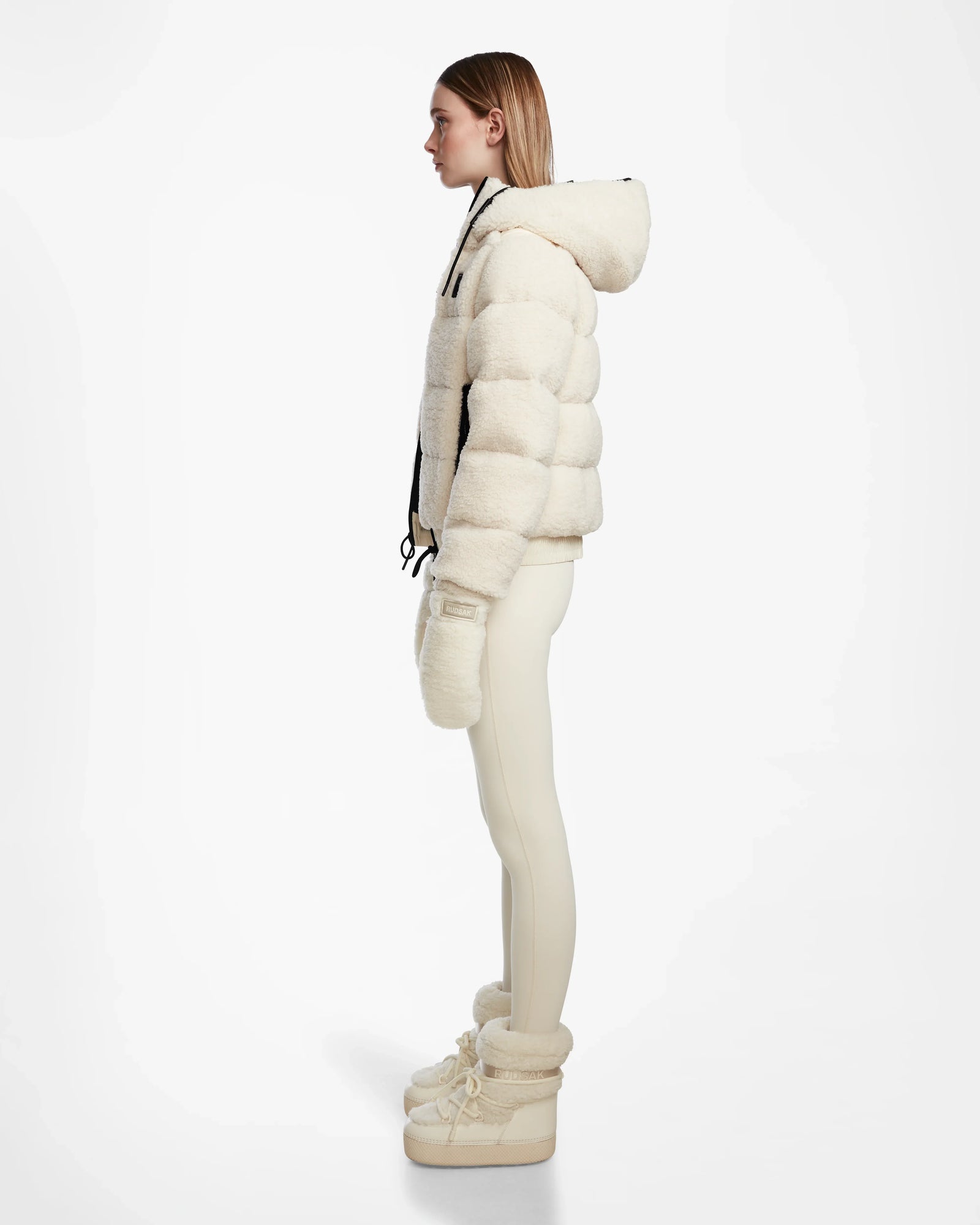 A woman stands in profile against a white background, wearing the DANI S - 8124537 Iceland cream cropped ski jacket by Rudsak, paired with matching mittens, fitted leggings, and fur-trimmed winter boots.