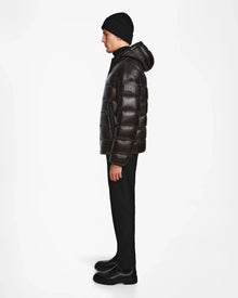 A man stands in profile against a white background, wearing the Rudsak DEMIAN S - M123508 Black men’s down puffer jacket, paired with black pants, black boots, and a black knit beanie.