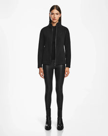 A woman faces forward against a white background, wearing the Rudsak EDITH - 8524617 Black hooded jacket with vegan leather trim, shiny black leggings, and black ankle boots.