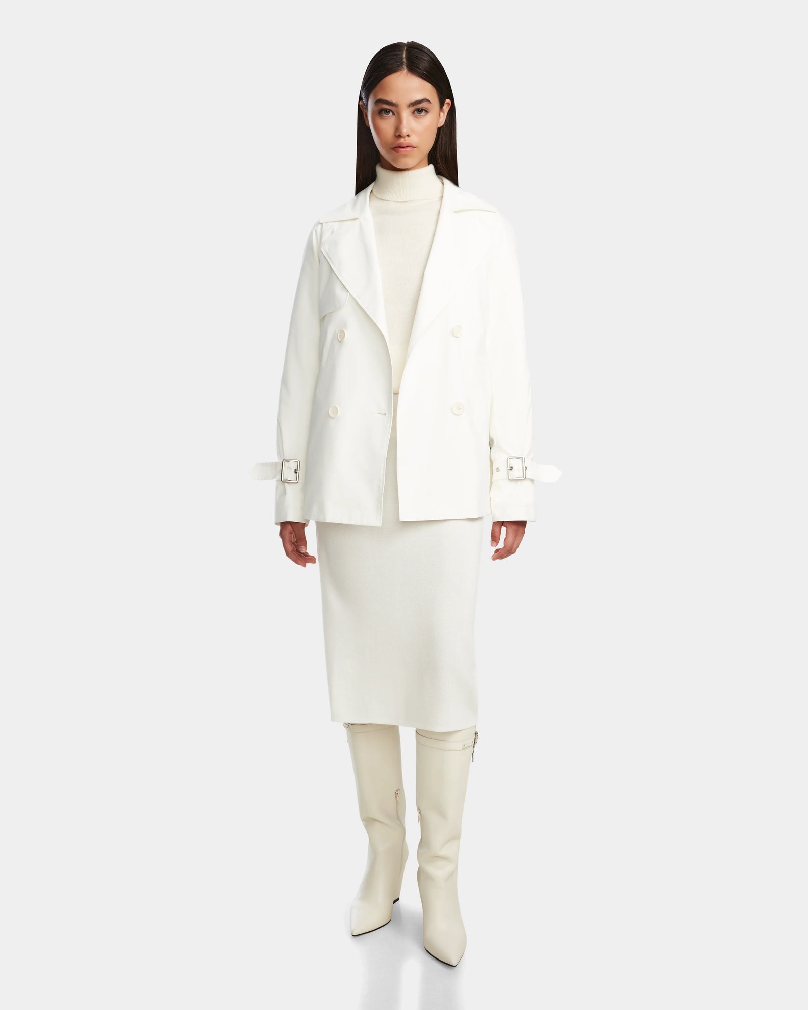 A woman stands facing forward in the Rudsak ELENOR - 8124707 Iceland white trench coat with a shearling collar, white turtleneck, knee-length skirt, and knee-high boots against a plain light background.