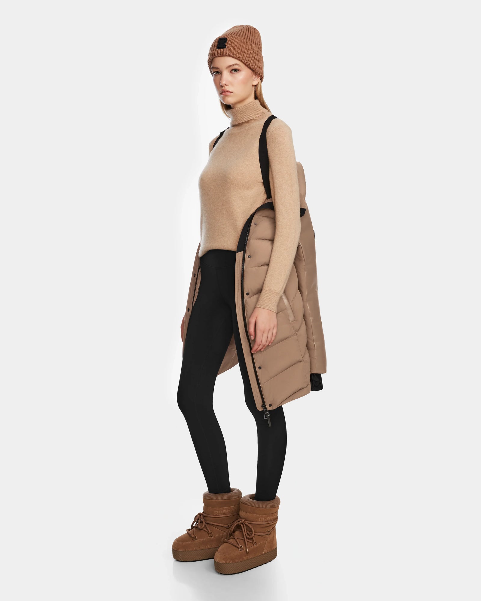A woman in a tan beanie, sleeveless turtleneck sweater, black leggings, and Rudsak ELSA - L123535 Sand RDS-certified down puffer coat draped over her shoulders stands in tan winter boots against a white background.