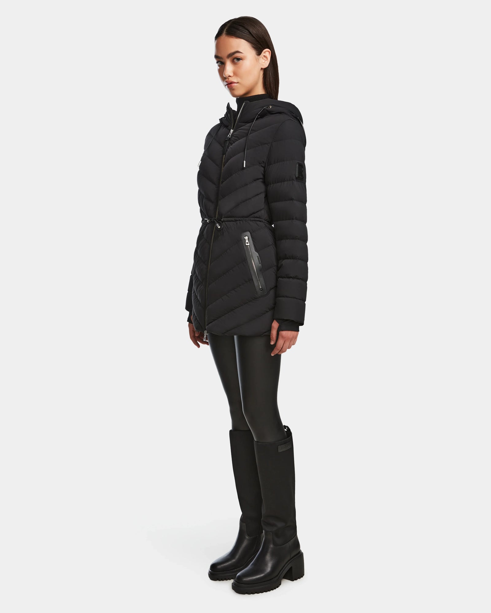A woman stands in profile wearing the Rudsak FRANCINE - 8124722 Black light down puffer jacket with a hood, black leggings, and knee-high black boots against a plain white background.