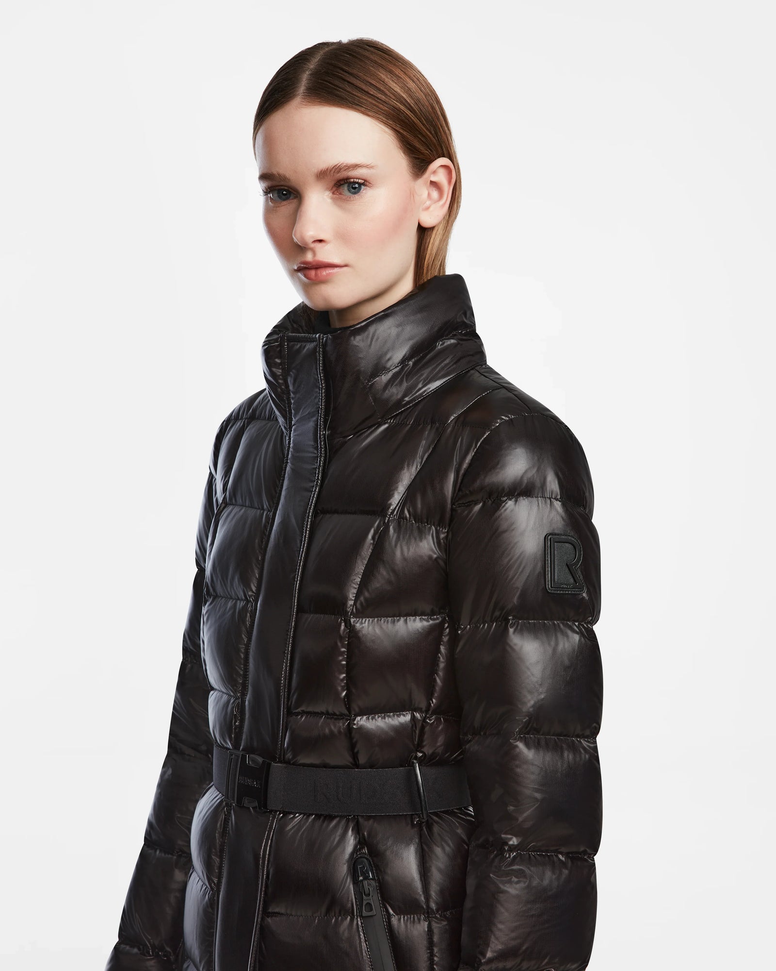 A woman with straight, light brown hair models the Rudsak FRIDA L-S - L124525 Black belted puffer coat, featuring a high collar and sleeve patch. This chic outerwear staple stands out against a plain white background.