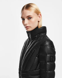 A woman with straight blonde hair models the Rudsak HANNA X - 8123558 Black women’s leather down jacket, showcasing its black quilted high-collar design and R logo on the sleeve, against a plain white background.