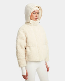 A woman in the Rudsak IKSA - 8124706 Iceland off-white teddy down puffer jacket with a matching hood and white pants stands against a plain background, looking slightly to the side with a neutral expression.