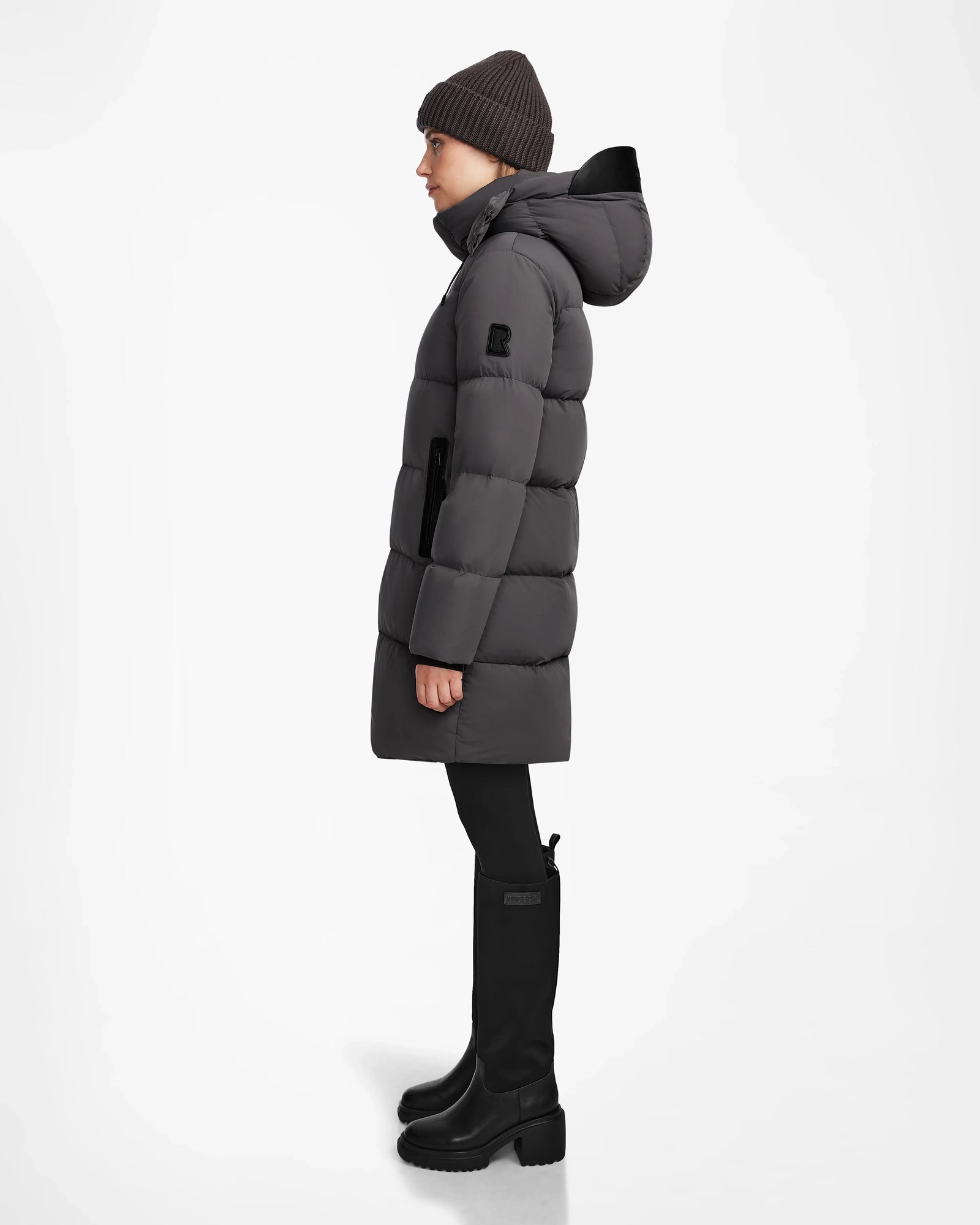 A person in a grey JOELLE - 8124729 puffer coat by Rudsak, black pants, knee-high boots, and a dark knit beanie stands in profile against a plain white background.