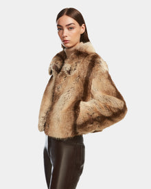 A woman with straight brown hair models the Rudsak JOSILYN - 8124792 Mocha women’s faux fur jacket in brown and beige, its plush texture paired with black leather pants as she poses against a plain white background.