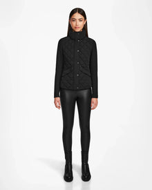 A woman with straight brown hair wears the Rudsak KARMA - L124503 Black matte light jacket with black leggings and boots, standing against a plain white background—ideal outerwear for transitional weather.